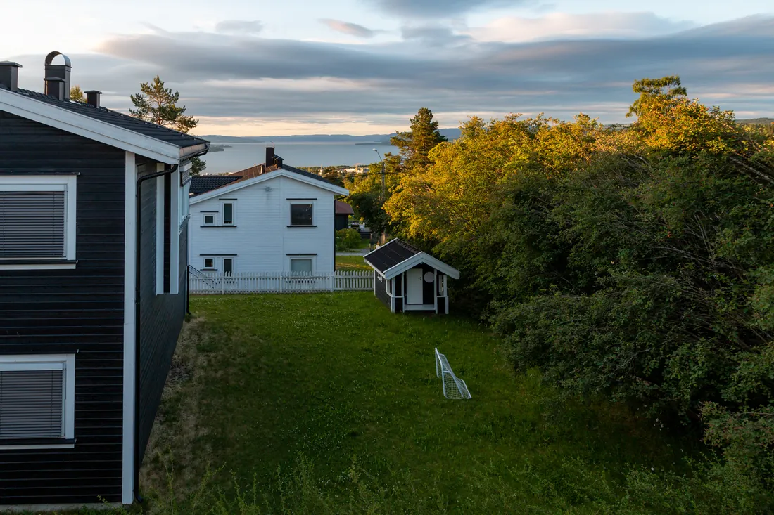 To vanlige norske eneboliger i mørkbeiset og hvitmalt tre, med hage, lekehus og utsikt mot vann i kveldslys