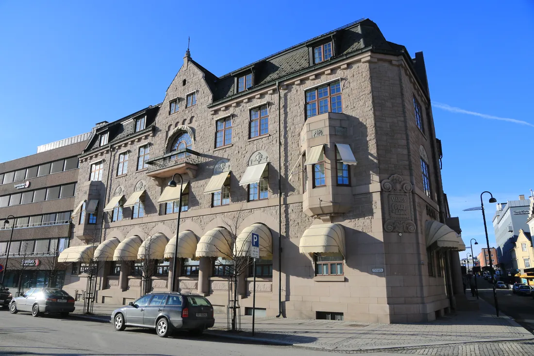 Hjørnet av et eldre bankbygg i grå naturstein i Hamar, med markiser over vinduene i første etasje og klar blå himmel over