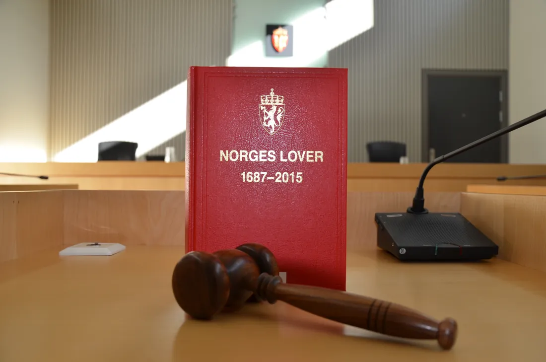 Lovboken Norges Lover stående på dommerbordet i en norsk rettssal, med klubbe foran og riksvåpenet på veggen bak