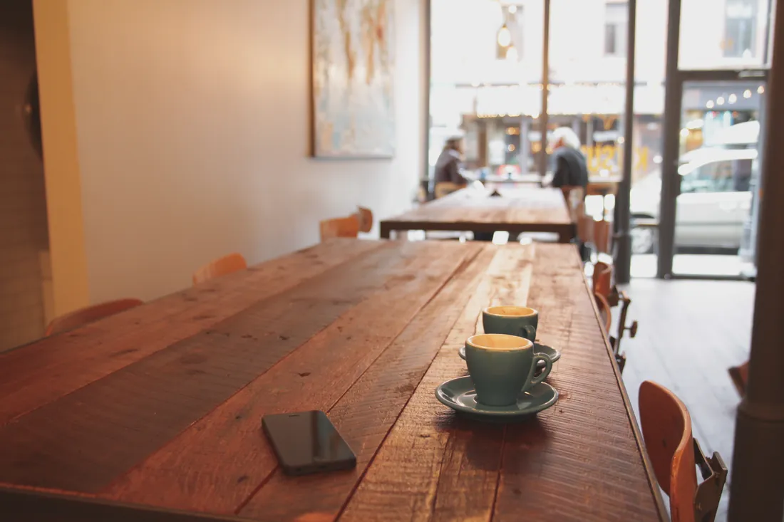 To små kaffekopper står ved siden av hverandre på et langt, slitt trebord i et lyst lokale med store vinduer