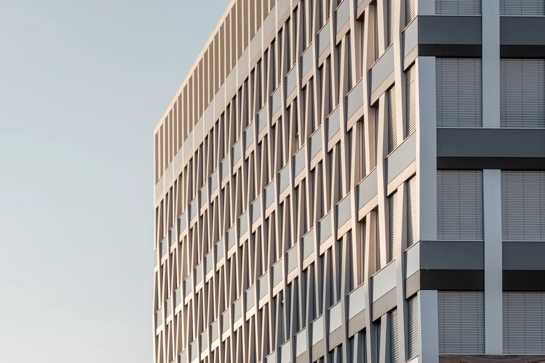 Hjørnet av et moderne kontorbygg med rutemønstret fasade i lyst betong og glass, mot en ren himmel i ettermiddagslys